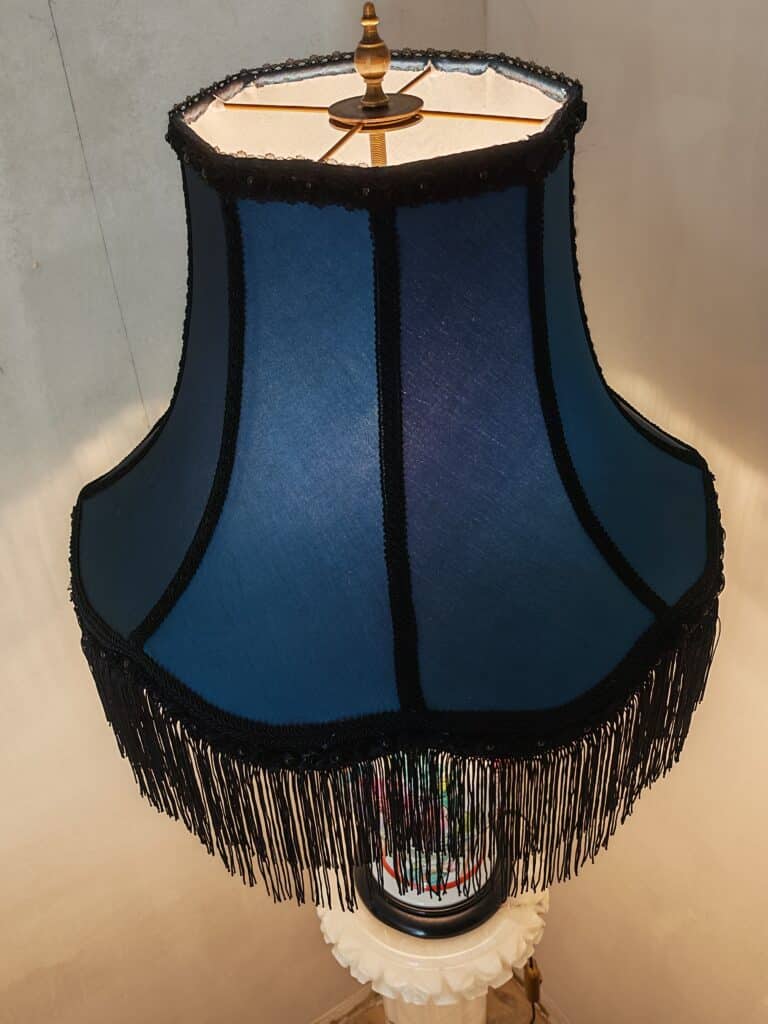 Abat-jour bleu évasé avec franges noires décoratives et tresses, sur une lampe vintage allumée. Éclairage chaleureux.