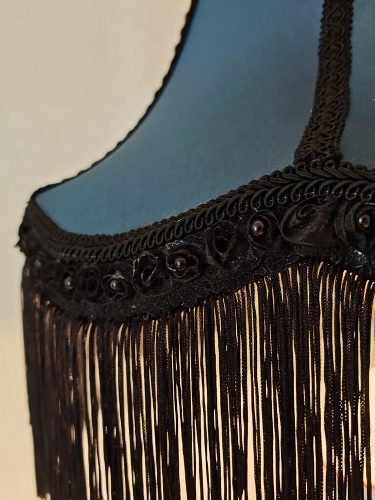 Détail abat-jour bleu-gris: passementerie noire, franges, roses en tissu et perles. Lumière chaude diffuse.