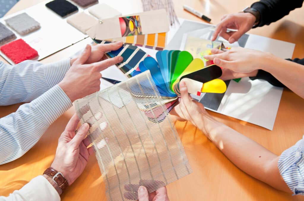 Quatre personnes collaborent autour d'une table, examinant des échantillons de tissus, palettes de couleurs et treillis métallique pour un projet de design.
