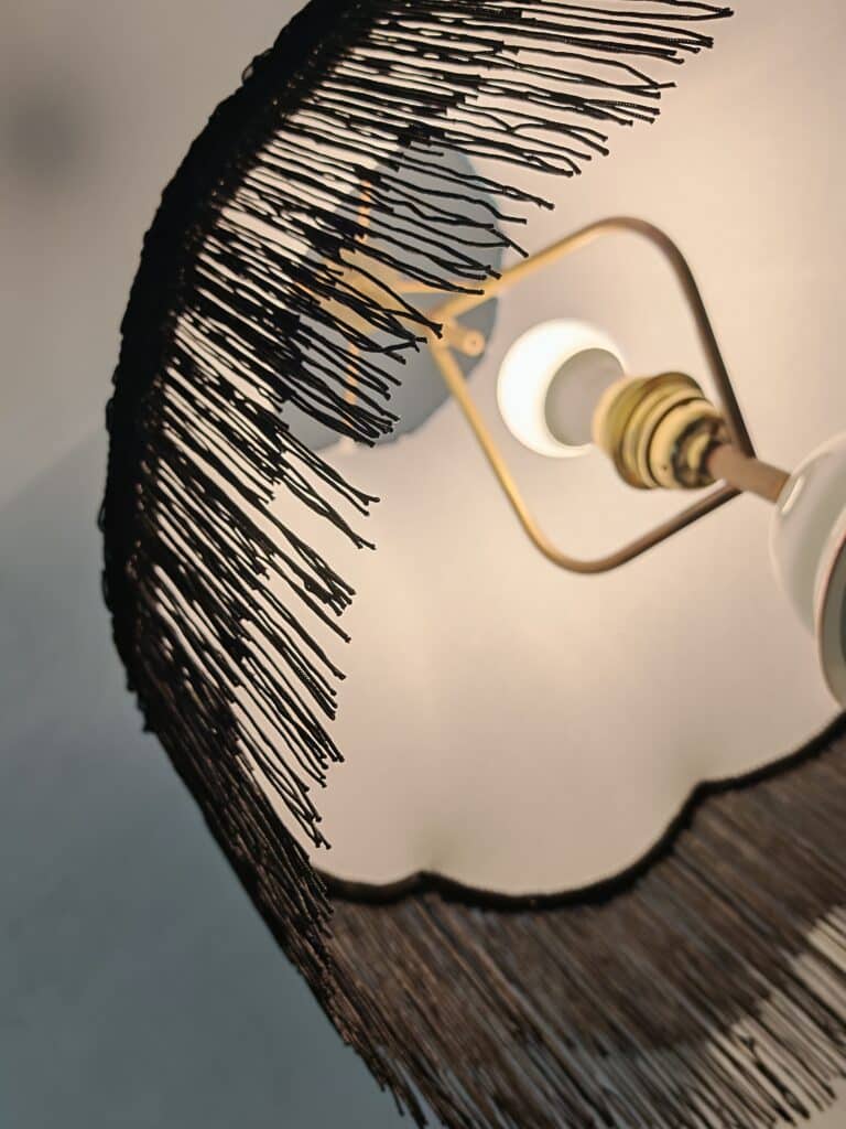 Lampe de plafond avec abat-jour à franges noires, révélant une ampoule allumée. Lumière chaude, design intérieur rétro-chic.